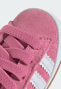 Růžové semišové tenisky s texturovanými tkaničkami, bílými akcenty a zigzagovým vzorem. Mají kulatou špičku a gumovou podrážku. Detailní pohled.