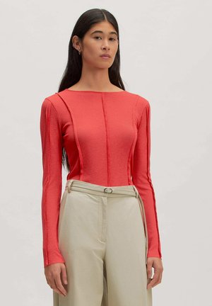Woman with long dark hair wearing a fitted coral long-sleeve top and high-waisted beige trousers with a belt, standing against a plain background.