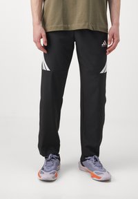 Man standing wearing black Adidas basketball pants with white stripes and gray sneakers with orange accents, hands relaxed by sides.