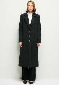 Cappotto lungo in misto lana grigio scuro con rever a punta, quattro bottoni frontali e finitura testurizzata. Abbinato a un top in maglia e pantaloni a gamba larga.