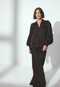 Blusa nera di satin con maniche a sbuffo e chiusura frontale con bottoni, abbinata a una lunga gonna nera. Tessuto liscio, silhouette aderente.