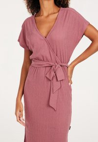 Robe portefeuille rose poussiére avec manches courtes, col en V, tissu texturé et ceinture nouée à la taille, dotée d'une fente latérale pour faciliter les mouvements.