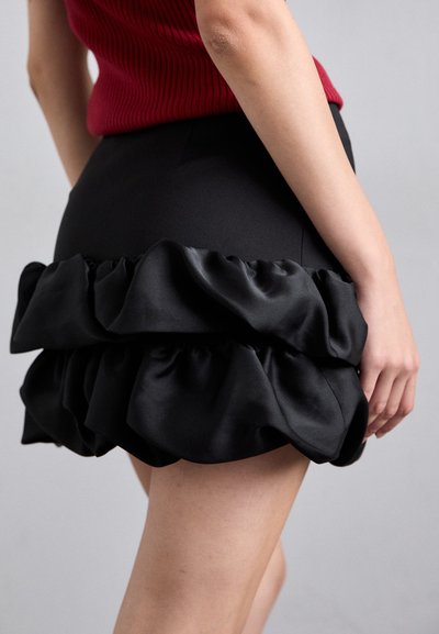 Femme portant une jupe mini noire avec des volants en satin superposés et un haut sans manches enroulé rouge, sur un fond uni.