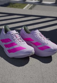 Par lyse lilla løbesko med klare pink striber placeret på tekstureret jord under sollys skygger.