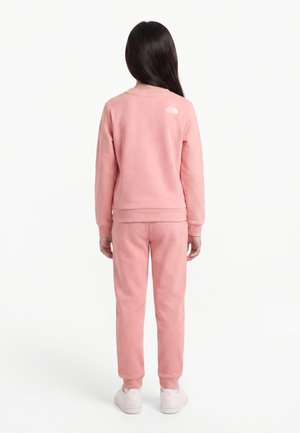 Ung person stående med ryggen til iført en pink sweatshirt og bukser med hvide sneakers på en hvid baggrund.
