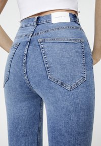 Jeans en denim bleu taille haute, avec un design ajusté, style à cinq poches et une patch en cuir blanc de marque à l'arrière.