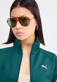 Giacca zip-up verde scuro con strisce Beige, caratterizzata da un piccolo logo bianco. Il modello indossa occhiali da sole marroni grandi e orecchini a cerchio.