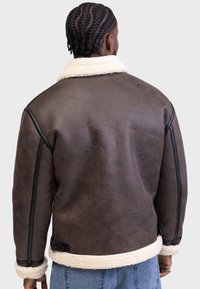 Bruine shearling jas met een witte fleece kraag, voorzien van zwarte accenten en gestructureerd leer. Inclusief zijriemen en een licht cropped ontwerp.