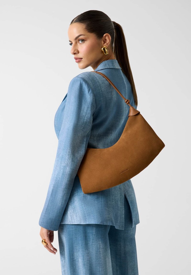 Femme avec une longue queue de cheval portant un costume bleu et des boucles d'oreilles en or, porte un sac à bandoulière en cuir marron, regardant sur le côté sur un fond uni.