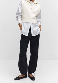 Ivoorkleurig gebreid vest met een hoge kraag over een wit overhemd met knopen, gecombineerd met losse zwarte cargobroeken en platte zwarte schoenen.