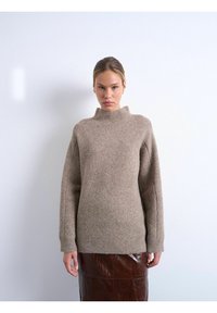 Femme aux cheveux lissés en arrière portant un pull beige en tricot avec un col montant et des manches longues, associé à une jupe en cuir marron foncé.
