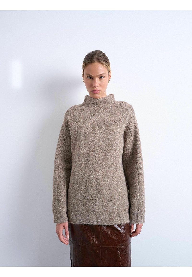 Femme aux cheveux lissés en arrière portant un pull beige en tricot avec un col montant et des manches longues, associé à une jupe en cuir marron foncé.