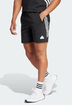 Mann trägt schwarze Adidas-Shorts mit weißen Streifen und Logo, kombiniert mit weißen Sneakers und schwarz-weiß gestreiften Socken.