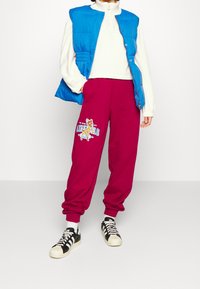 Person som bär en vit långärmad topp, en blå pufferväst, magentaröda träningsbyxor med en tecknad älg-illustration, vita strumpor och svart-vita sneakers.