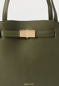 Olívazöld kézitáska sima textúrával, téglalap alakú arany kapoccsal és alul bemélyített logóval. Két fogóval ellátva.