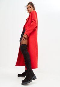 Un abrigo largo rojo hecho de un material suave, combinado con botas de ante negras alargadas y una falda negra, mostrando una silueta estilizada.