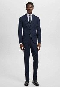 Marinblå kostym med en strukturerad design, med en tvåknappsblazer, raka byxor, vit skjorta och ett randigt slips. Glansiga svarta skor.
