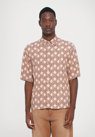 Un homme aux cheveux courts portant une chemise beige à boutons ornée de motifs de feuilles abstraites rouge foncé et un pantalon marron clair, se tenant contre un fond blanc.