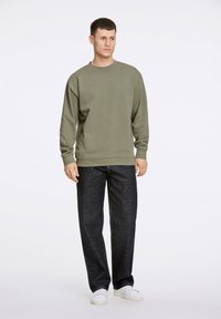 Sudadera verde olivo con cuello redondo y puños acanalados, combinada con pantalones de mezclilla rectos oscuros y zapatillas blancas, de pie sobre un fondo liso.