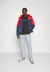 Giacca a vento rossa e blu navy, in cotone misto con zip, cappuccio e patch con logo. Abbinata a una felpa nera, jeans chiari e sneaker argentate.