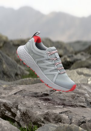 Chaussure de trail Columbia gris clair et corail en suspension au-dessus d'un terrain rocailleux avec un arrière-plan montagneux flou.