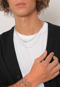 Sølvsmykke lagdelt over en hvid t-shirt, båret med en sort cardigan. Bemærkelsesværdige detaljer inkluderer en rektangulær vedhæng og et kædedesign.