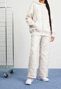 Ljust beige zip-up hoodie med framfickor, tillsammans med vita quiltade vida byxor med en diamantmönster, kompletterat med vita sneakers.