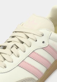 Λευκό δερμάτινο αθλητικό παπούτσι με τρεις ρίγες σε ανοιχτό ροζ, κορδόνια με υφή και μπεζ σόλα. Διαθέτει ανάγλυφο λογότυπο στη γλώσσα.