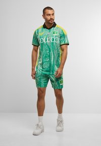 Mann steht mit einem grün gemusterten Sporttrikot und Shorts mit gelben Akzenten, weißen Socken und weißen Turnschuhen vor einem einfarbigen Hintergrund.