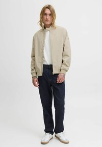 Veste bomber beige avec fermeture éclair, poignets côtelés, portée sur une chemise blanche. Un jean bleu marine et des baskets blanches complètent le look.
