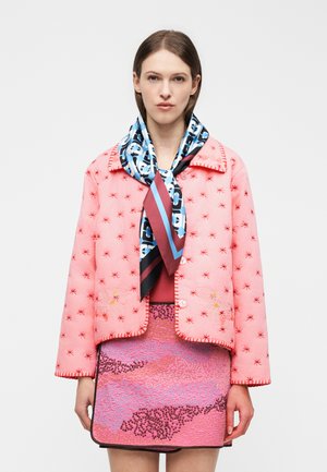 Kvinde iført en pink quiltet jakke med rødt stjernebroderi, mønstret pink nederdel og et blå, sort og mørkerødt mønstret tørklæde bundet om halsen.