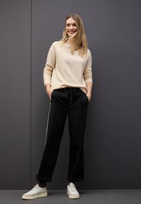 Beiger, langärmeliger Pullover kombiniert mit lockeren, schwarzen Sweatpants mit Kordelzug und seitlichen Streifen. Weiße Sneakers runden das Outfit ab.