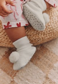 Chaussons en tricot gris clair avec un revers et un pompon duveteux à l'avant ; semelles en caoutchouc à picots pour une meilleure adhérence.