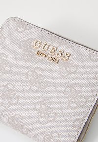 Lys grå teksturert lommebok med pregete mønstre og hardware i gulltone. Har glidelås og merkevarenavnet "GUESS" lenge vist.