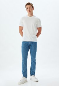T-shirt bianca in cotone con maniche corte, abbinata a jeans in denim blu chiaro. Il modello indossa sneakers bianche, in piedi contro uno sfondo chiaro.