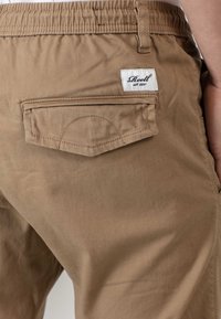 Bermudas de color marrón claro fabricadas en algodón, con un bolsillo trasero con tapa y una etiqueta blanca cosida con letras oscuras.