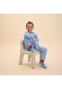 Blauwe gestreepte trainingsbroek met rode accenten en elastische manchetten, gedragen met lichtblauwe sneakers, zittend op een gestructureerde, gespikkelde kunststof kruk.