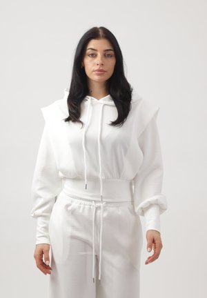 Vrouw met lang donker haar draagt een witte hoodie met opgeblazen mouwen en bijpassende witte joggingbroek tegen een effen lichte achtergrond.