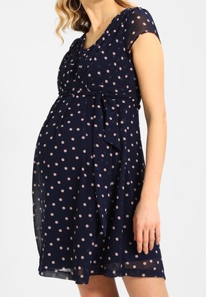 Kobieta w ciąży ubrana w granatową sukienkę z krótkim rękawem w jasnoróżowe kropki, z paskiem w talii, stoi na białym tle.