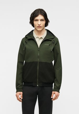 Jeune personne aux cheveux mi-longs foncés portant une veste zippée verte et noire par-dessus une chemise beige à col, se tenant devant un fond uni.