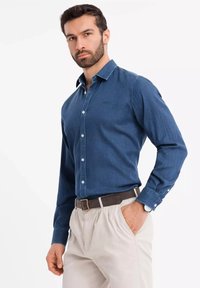 Camicia di jeans blu scuro con colletto button-down, maniche lunghe e bottoni azzurri. Abbinata a pantaloni beige e una cintura marrone.