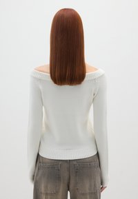 Schulterfreier weißer Pullover mit gerippten Bündchen und Saum, kombiniert mit grauen, leicht verwaschenen, hochgeschnittenen Hosen. Glatte Textur.