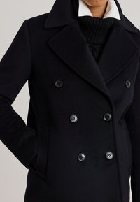 Czarny dwurzędowy wełniany płaszcz z dużymi klapami, dwoma bocznymi kieszeniami i czarnymi guzikami, noszony na czarnym dzianinowym golfie.