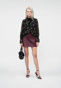 Donna che indossa una blusa nera trasparente a fiori, una mini gonna in pelle bordeaux, sandali neri con tacco alto e tiene in mano una borsa nera.