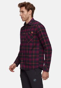 Uomo che indossa una camicia a quadri rossa e nera con tasche sul petto e pantaloni neri con un logo blu sulla coscia, in piedi di lato.