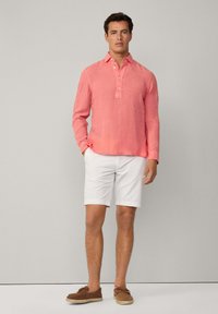 Camicia a maniche lunghe in lino corallo con pattina dei bottoni, abbinata a un pantalone corto bianco e calzature marroni. Design semplice, tessuto leggero.