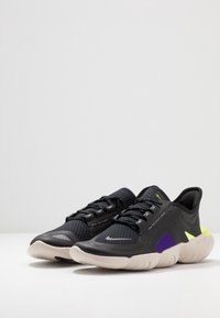 Un paio di scarpe da atletica nere con suole bianche, dettagli in viola e giallo neon, tomaia in rete e chiusura con lacci su una superficie bianca.