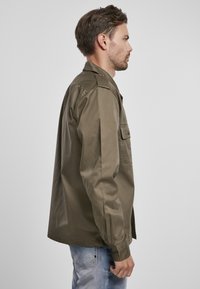 Camisa de manga larga color verde oliva, con un bolsillo frontal, puños abotonados y un diseño recto, confeccionada en un material suave.