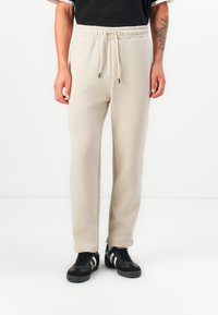 Beige sweatpants with a drawstring waist, tapered legs, and smooth texture. Paired with black sneakers featuring white stripes.