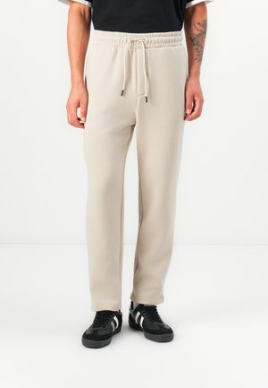 Beige sweatpants with a drawstring waist, tapered legs, and smooth texture. Paired with black sneakers featuring white stripes.